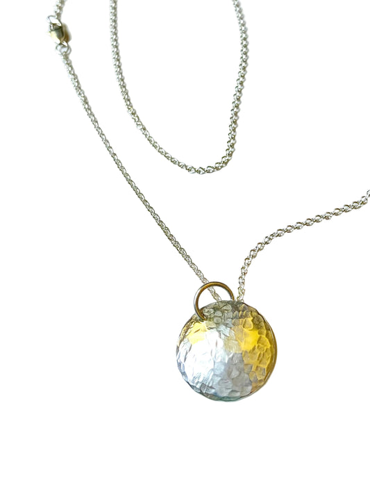 A hammered round pendant necklace with a textured silver disc and a ring attachment, suspended from a delicate silver chain on a white background.