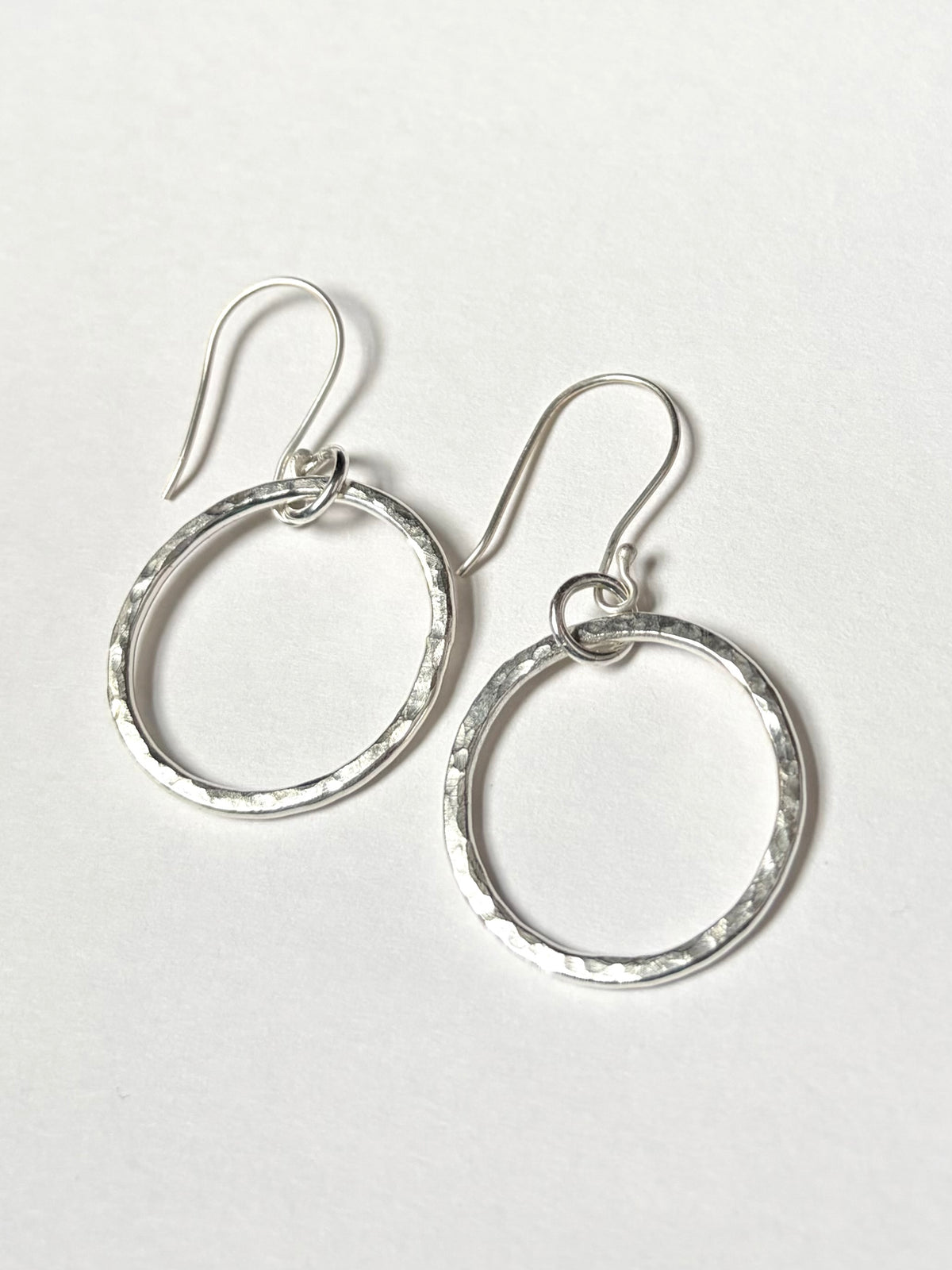 A pair of silver open hoop dangle earrings with a hammered texture and fishhook backings, displayed on a clean white background.
