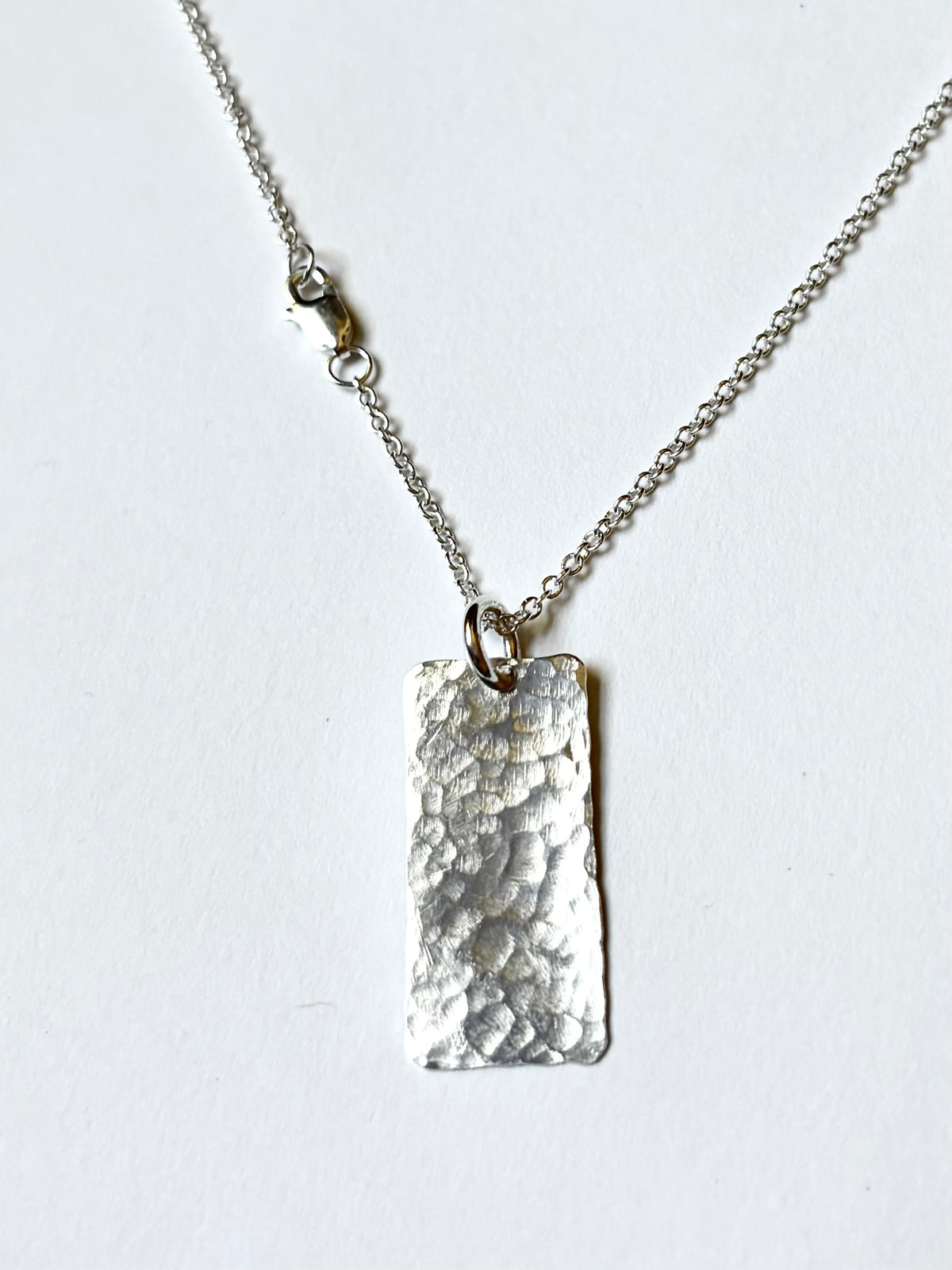 A silver necklace featuring a hammered rectangular pendant on a delicate silver chain, photographed against a plain white background.