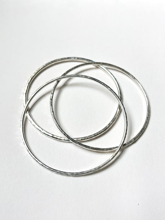 Three thin silver metal bangles with a hammered texture, overlapping on a plain white background.