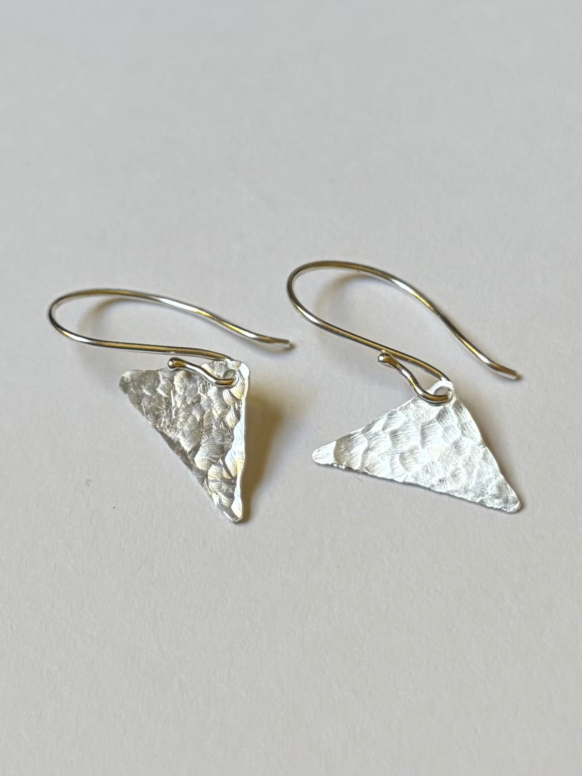 A pair of hammered triangular silver dangle earrings with fishhook wires, displayed on a light background.
