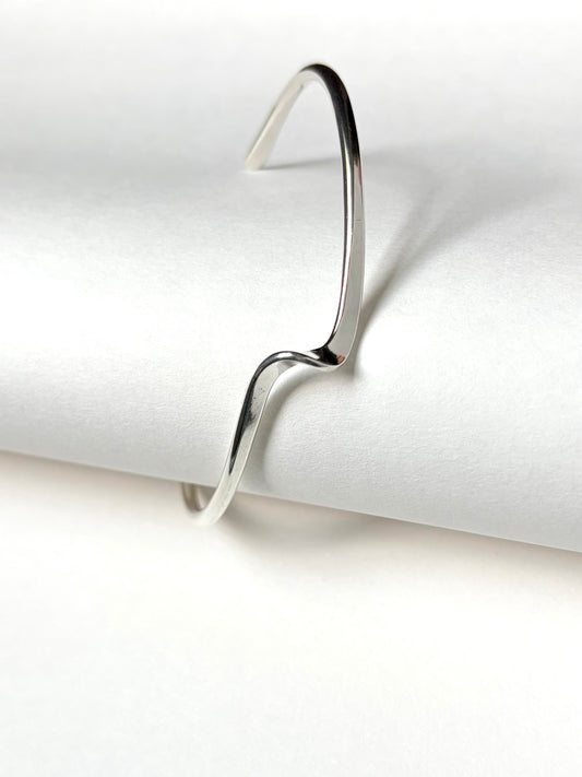 A minimalist silver open cuff bracelet with a wavy, fluid line design, displayed on a clean white textured background.