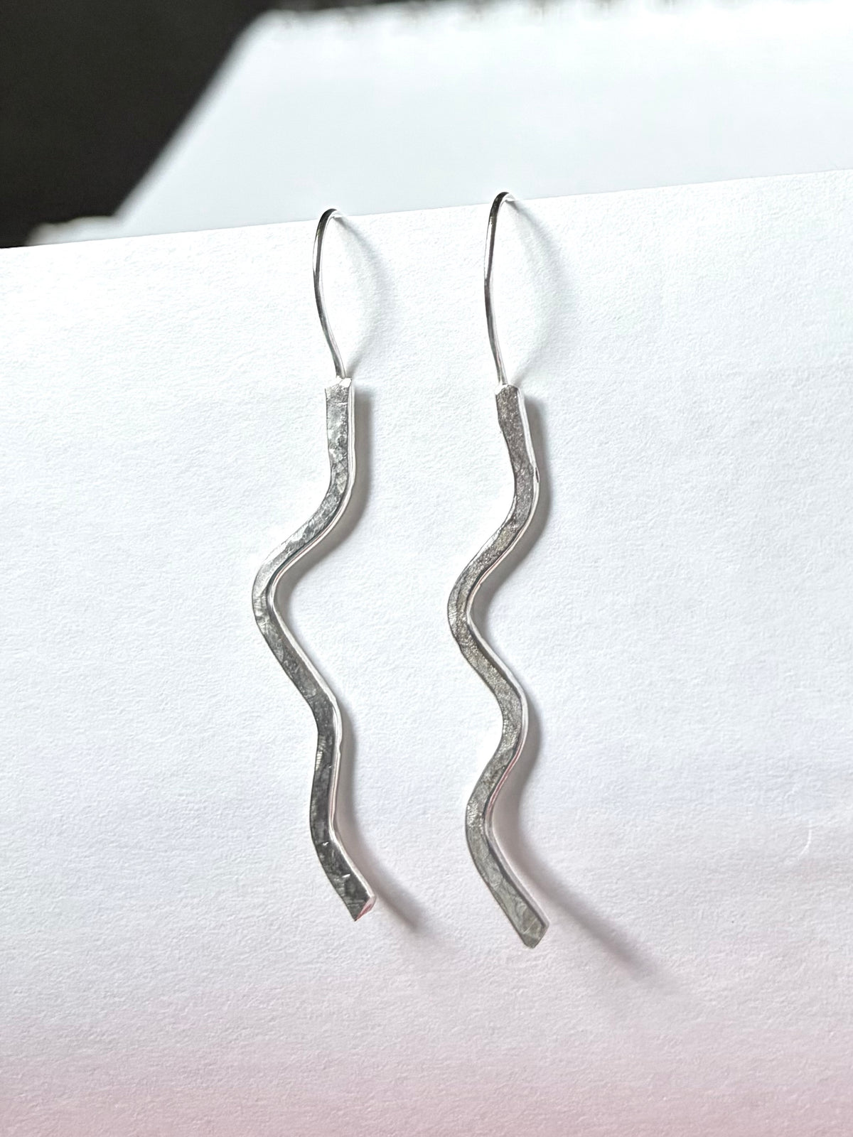 A pair of silver wavy drop earrings with textured, hammered finishes and simple fishhook wires, displayed against a white paper background.