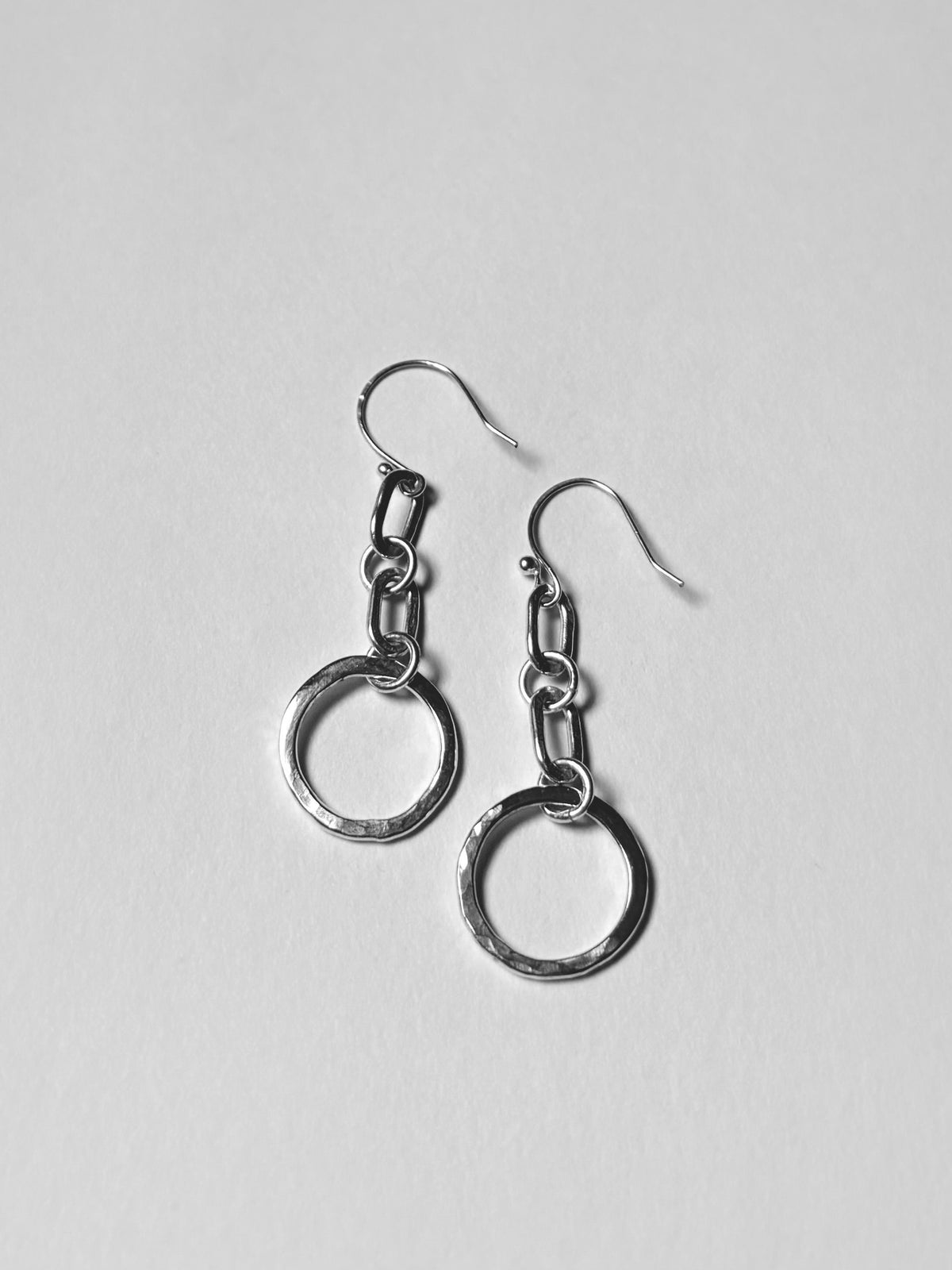 A pair of silver drop earrings with textured circular rings and chain links, displayed on a light gray background.