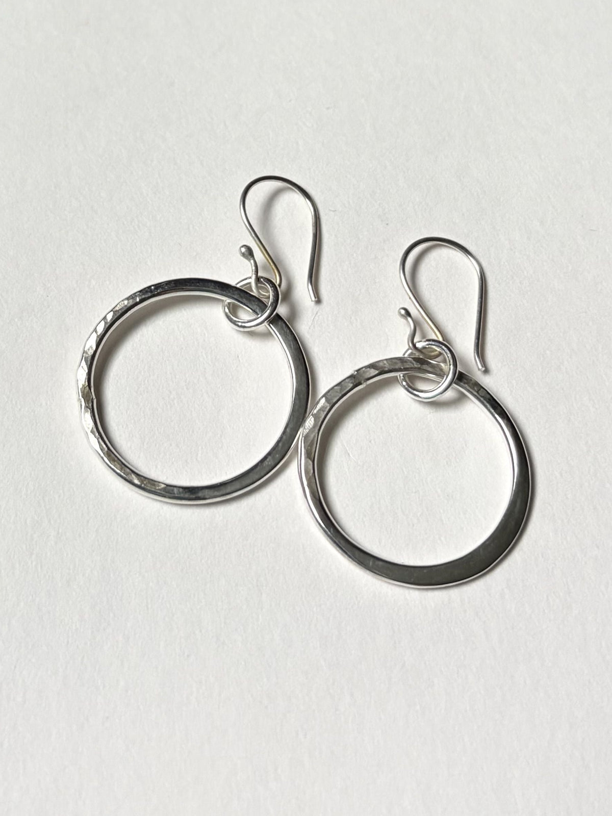 A pair of silver-toned open hoop earrings with fishhook backings, displayed on a clean, light off-white textured background.
