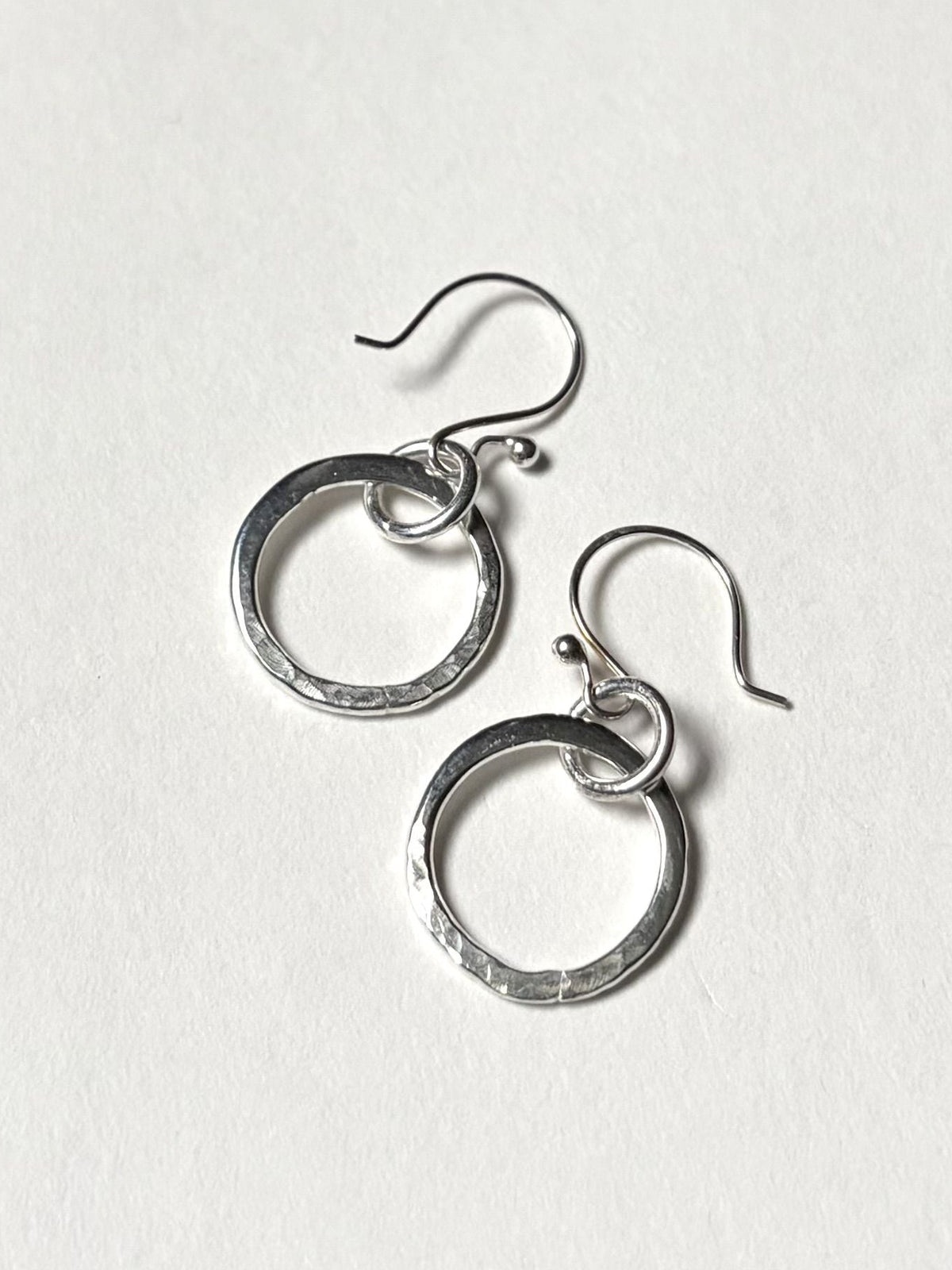 A pair of silver dangle earrings with textured circular hoops and fishhook wires, displayed on a clean, off-white textured background.