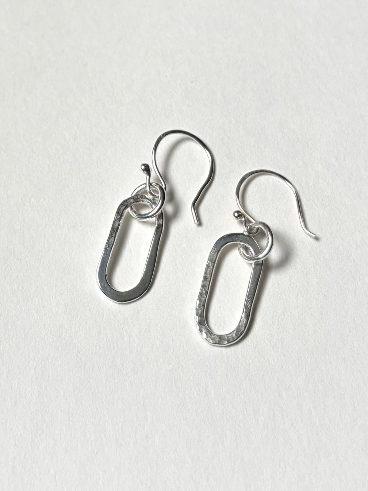 A pair of silver drop earrings with textured oval links and fishhook backings, displayed on a clean, light off-white textured surface.