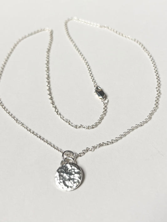 A silver chain necklace with a textured, hammered circular pendant, displayed on a clean white background.