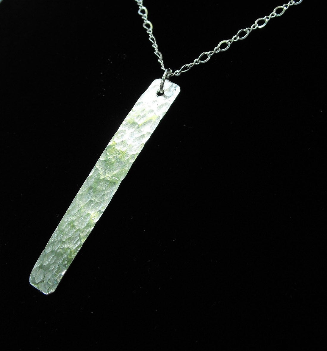 A long, flat, thin, shiny hammered silver pendant of approximately.5 inch by 1.75 inches in size is hanging from a small silver jump ring, which is hanging from a silver chain.