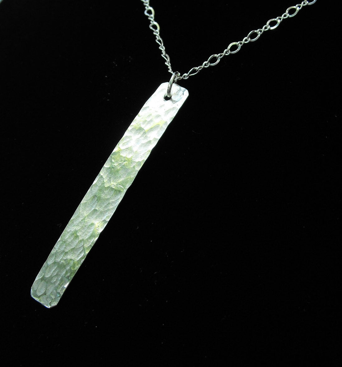 A long, flat, thin, shiny hammered silver pendant of approximately.5 inch by 1.75 inches in size is hanging from a small silver jump ring, which is hanging from a silver chain.