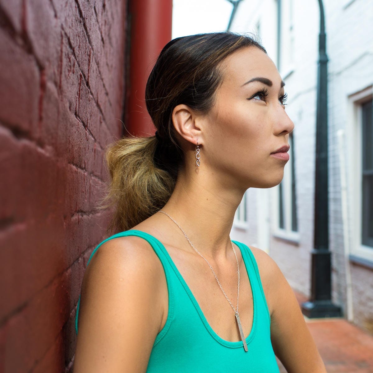 A woman in a green tank top and brown hair pulled into a pony tail is gazing upward.  She is wearing a long flat rectangular silver pendant on a silver chain. She is also wearing silver dangle earrings.