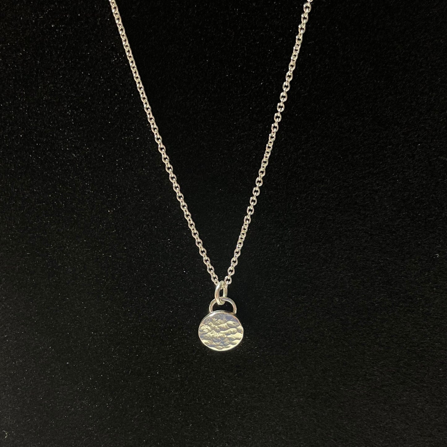 May include: Silver necklace with a hammered, circular pendant. The chain is delicate and the pendant is textured, adding a rustic touch. This minimalist necklace is perfect for everyday wear or a special occasion. The simple design makes it a versatile piece that can be styled in many ways.