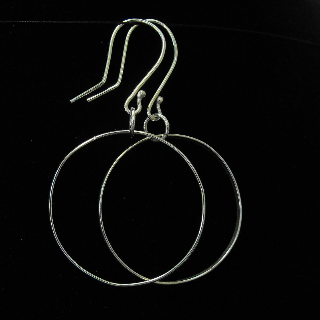 May include: A pair of silver-toned hoop earrings featuring thin, circular wire frames suspended from curved ear wires. The earrings have a polished metallic finish and a simple, open-circle design that hangs vertically from the hook attachments.