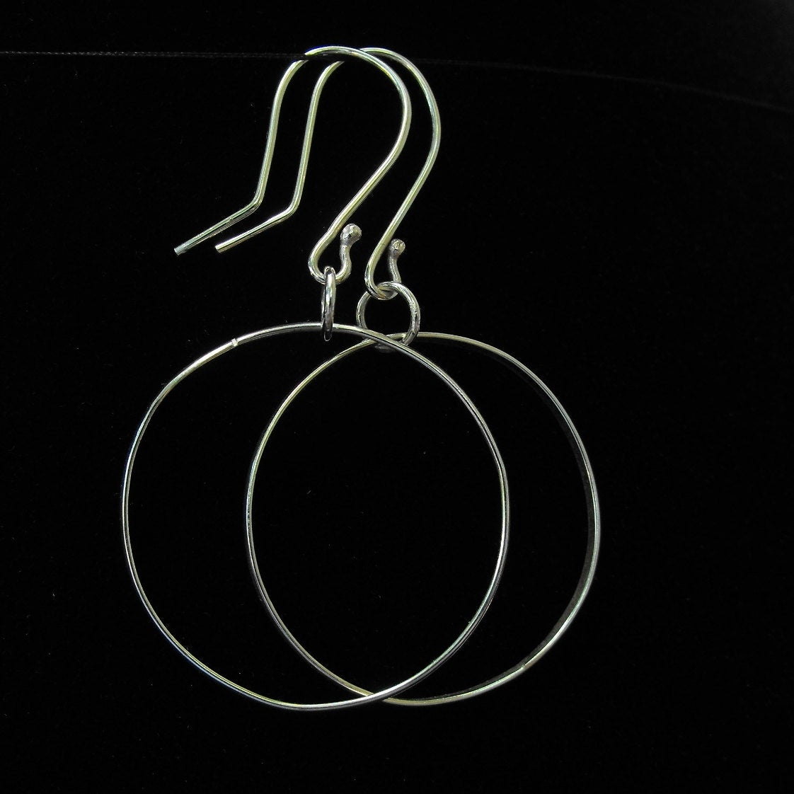May include: A pair of silver-toned hoop earrings featuring thin, circular wire frames suspended from curved ear wires. The earrings have a polished metallic finish and a simple, open-circle design that hangs vertically from the hook attachments.