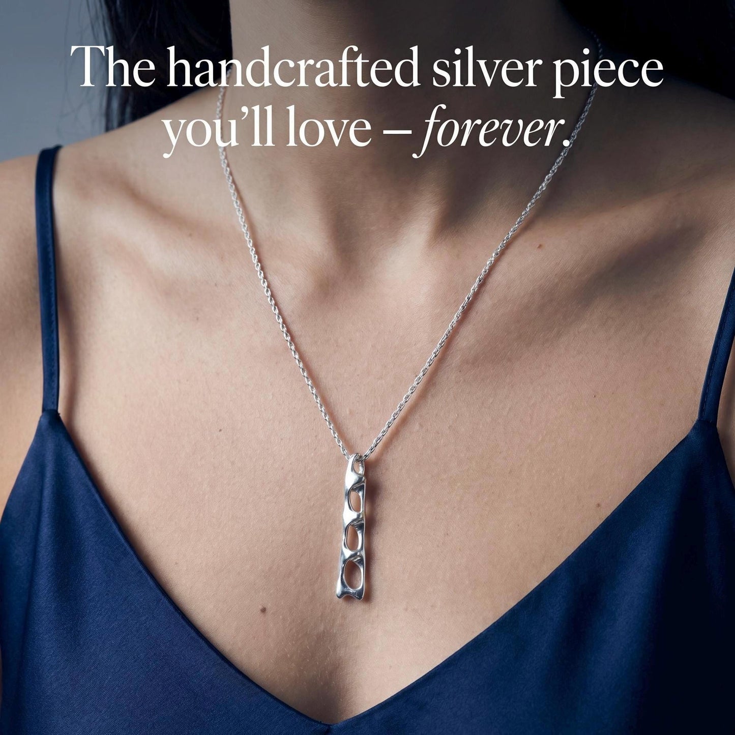 May include: A silver necklace featuring a long, vertical pendant with an openwork, abstract link design. The text "The handcrafted silver piece you'll love – forever." is overlaid on the image. The pendant hangs from a thin, textured silver chain.
