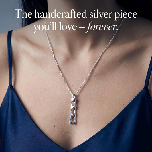 May include: A silver necklace featuring a long, vertical pendant with an openwork, abstract link design. The text "The handcrafted silver piece you'll love – forever." is overlaid on the image. The pendant hangs from a thin, textured silver chain.