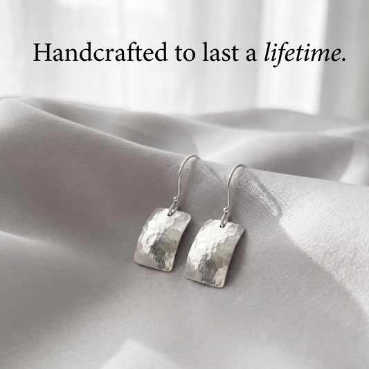 May include: A pair of silver drop earrings featuring rectangular, hammered metal pendants with curved edges and hook-style findings. The text "Handcrafted to last a lifetime." is displayed above the earrings in a black serif font.