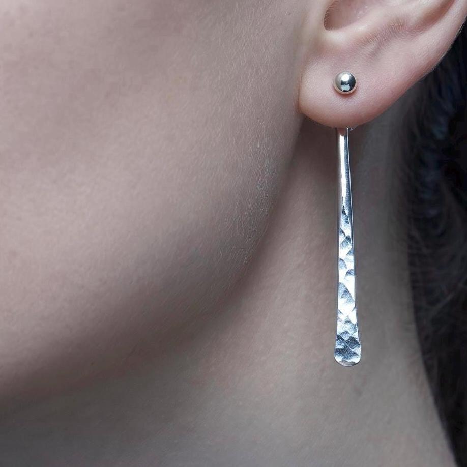 May include: A silver earring featuring a small spherical stud at the earlobe with a long, slender, hammered-texture drop attachment. The text MODERN ESSENTIALS ARGENTIUM SILVER is overlaid on the image in white, sans-serif and serif fonts.
