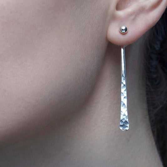 May include: A silver earring featuring a small spherical stud at the earlobe with a long, slender, hammered-texture drop attachment. The text MODERN ESSENTIALS ARGENTIUM SILVER is overlaid on the image in white, sans-serif and serif fonts.