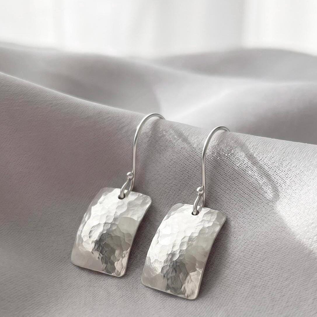 May include: A pair of silver drop earrings featuring rectangular, hammered metal pendants with curved edges and hook-style findings. The text "Handcrafted to last a lifetime." is displayed above the earrings in a black serif font.