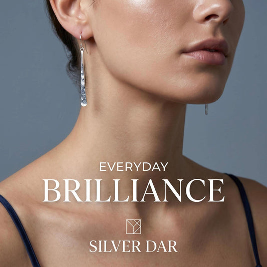 May include: A pair of long, slender, silver drop earrings with a hammered, textured finish. The earrings hang from thin silver hooks. The image includes the text "EVERYDAY BRILLIANCE" and "SILVER DAR" along with a small geometric logo.