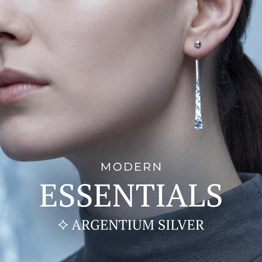 May include: A silver earring featuring a small spherical stud at the earlobe with a long, slender, hammered-texture drop attachment. The text MODERN ESSENTIALS ARGENTIUM SILVER is overlaid on the image in white, sans-serif and serif fonts.