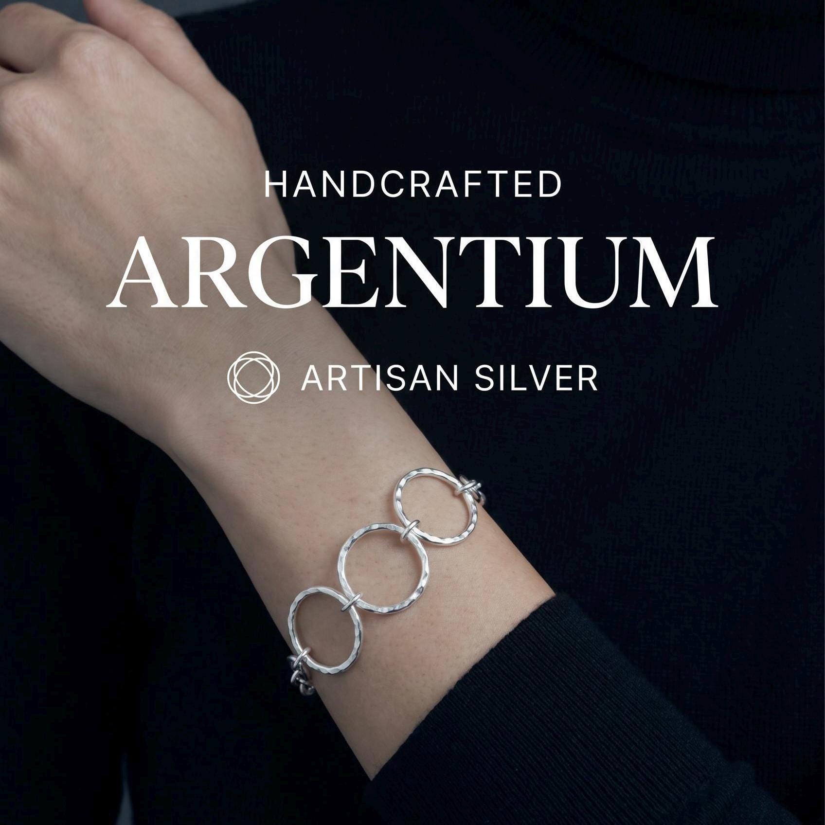 May include: A silver bracelet featuring three interconnected circular links with a hammered, textured finish. The text HANDCRAFTED ARGENTIUM ARTISAN SILVER is displayed above the bracelet, along with a circular geometric logo icon.