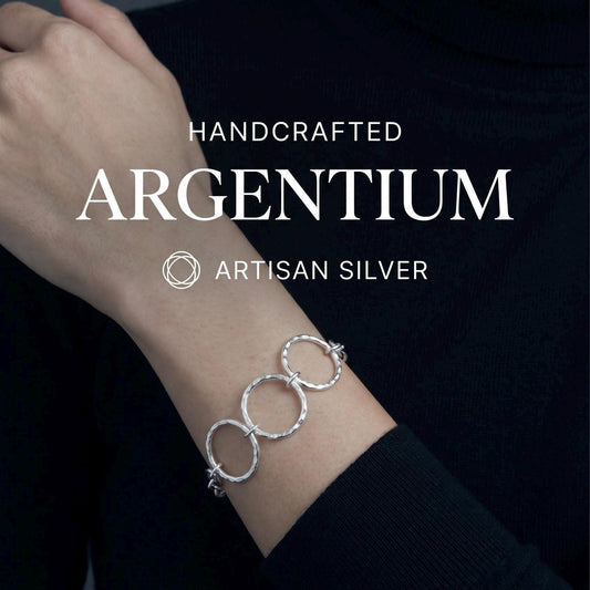May include: A silver bracelet featuring three interconnected circular links with a hammered, textured finish. The text HANDCRAFTED ARGENTIUM ARTISAN SILVER is displayed above the bracelet, along with a circular geometric logo icon.