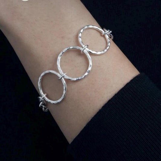 May include: A silver bracelet featuring three interconnected circular links with a hammered, textured finish. The text HANDCRAFTED ARGENTIUM ARTISAN SILVER is displayed above the bracelet, along with a circular geometric logo icon.