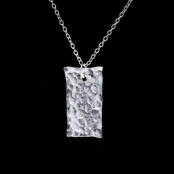 A flat, shiny, silver rectangle of .5 inch by 1 inch pendant with a top drilled hole for a silver jump ring dangles from a curb chain.  The pendant has been hammered and has slightly uneven edges.